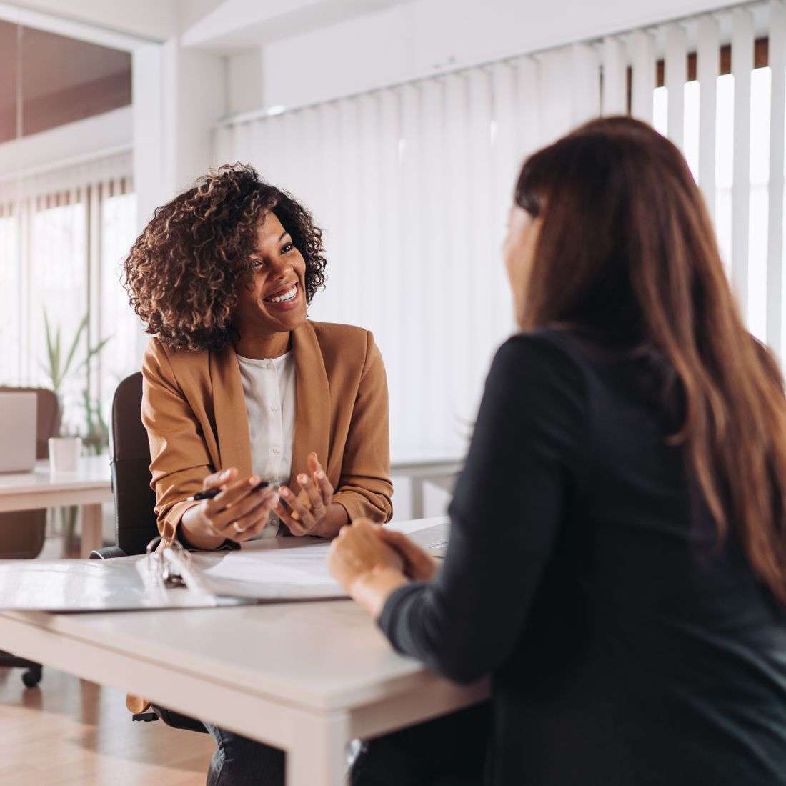 A smiling Black consultant in a camel blazer meeting with a client across a desk