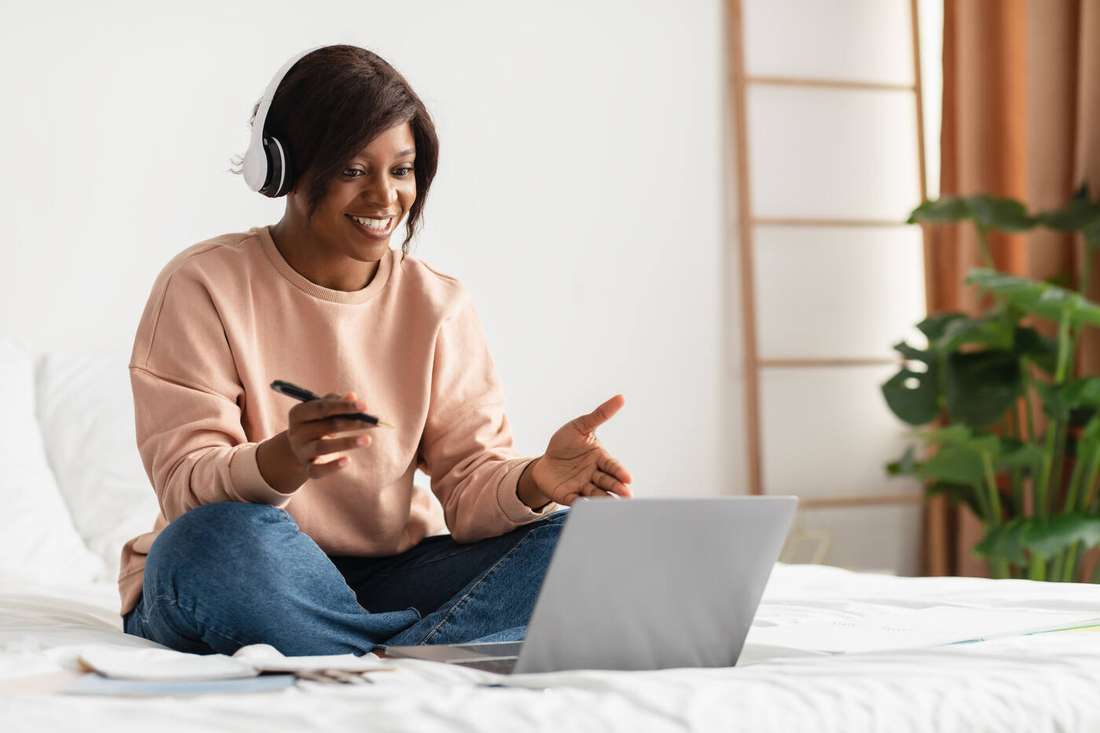 A smiling woman sitting on her bed with notes and a laptop, learning from home