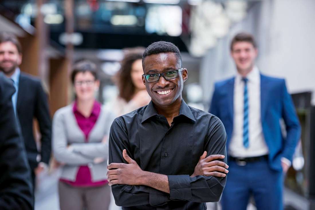 A smiling young Black man in glasses with arms crossed, colleagues standing behind him in a busy office