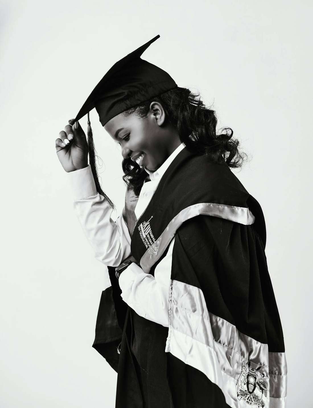 A young Black girl in a graduation cap and gown, adjusting her tassel, in black and white