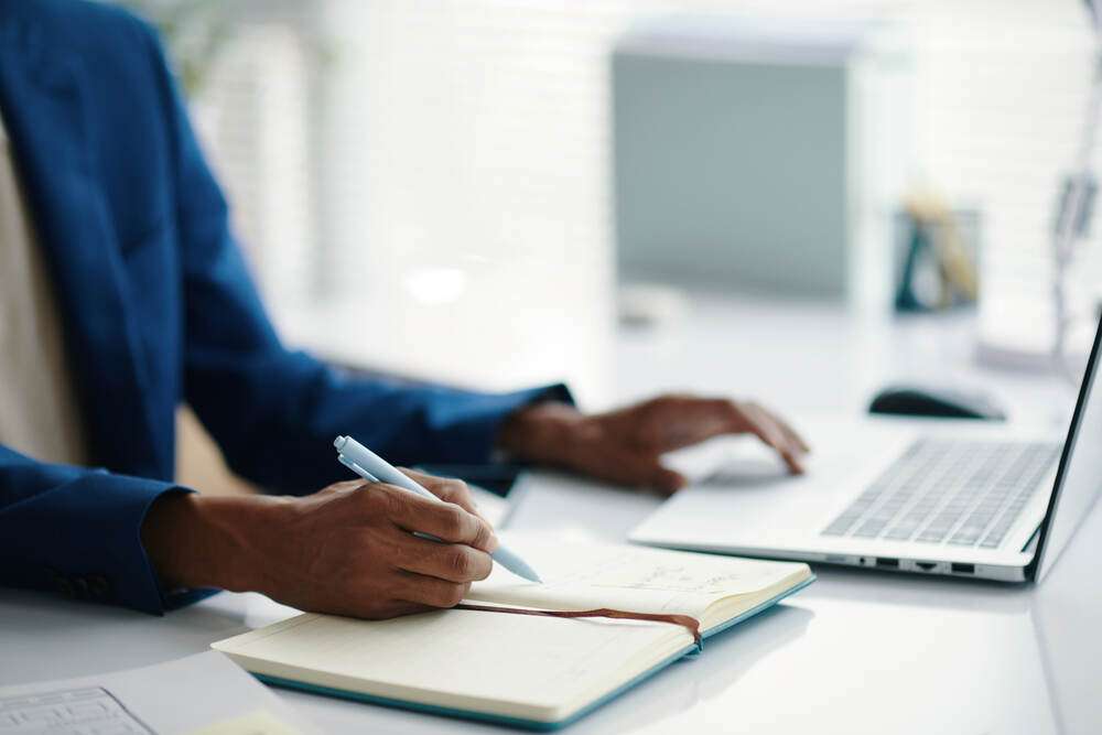Hands writing in a notebook beside a laptop
