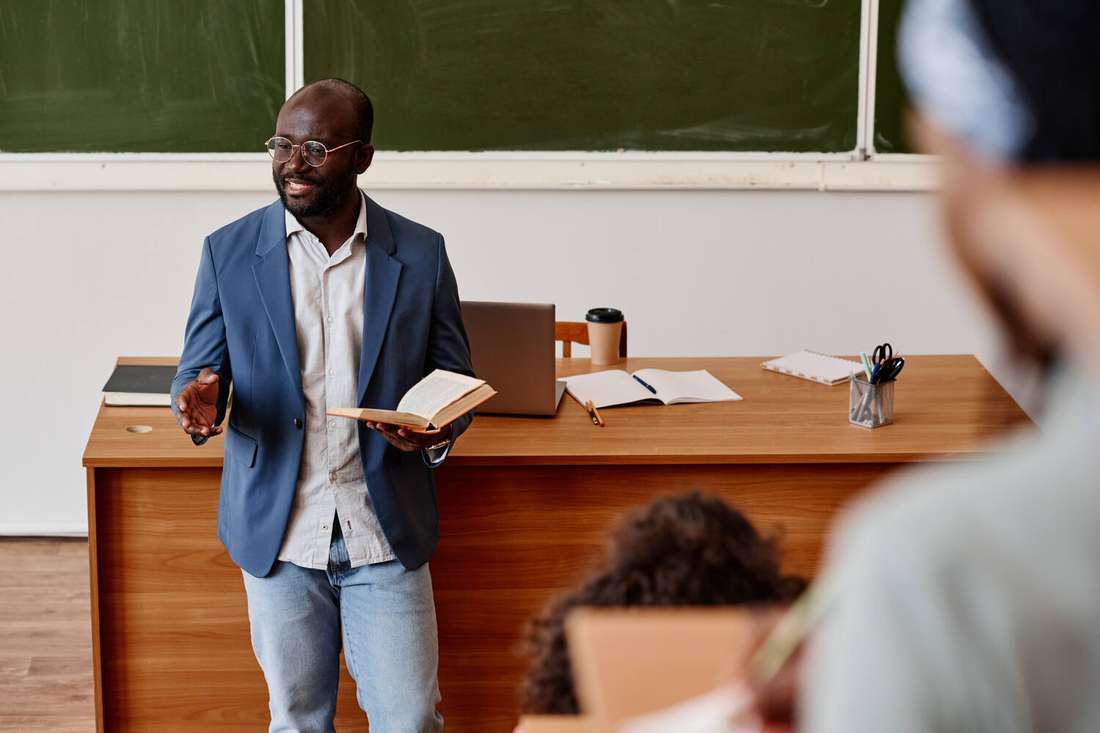 A Black male teacher holding a book, speaking to a classroom