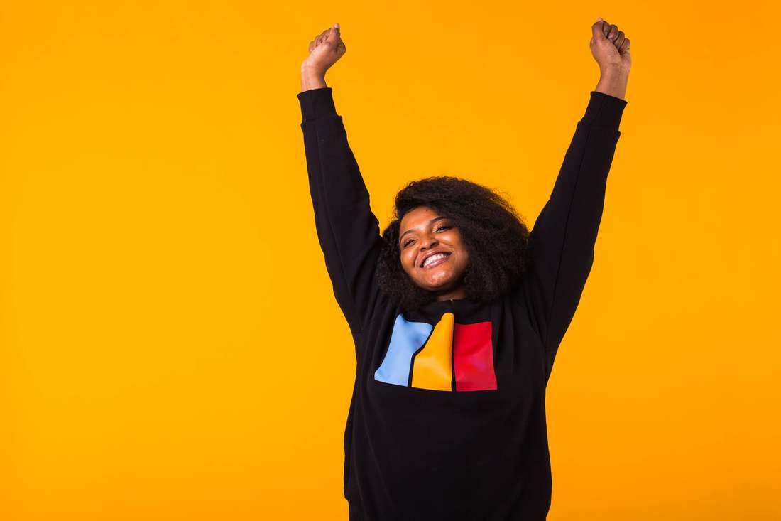 A joyful Black young woman with arms raised against a bright yellow background