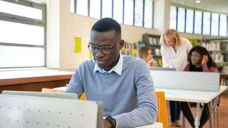 A young Black man in glasses working on a laptop in a school library