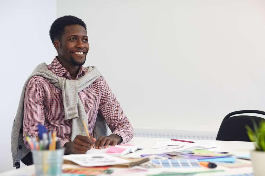 A smiling young man at a desk with art and writing supplies