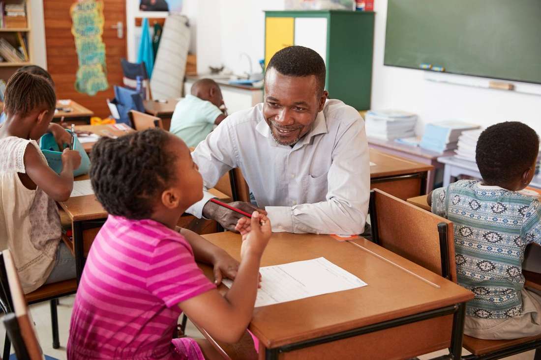 A smiling Black male teacher sitting with a young girl at her desk, helping with her work in a busy classroom