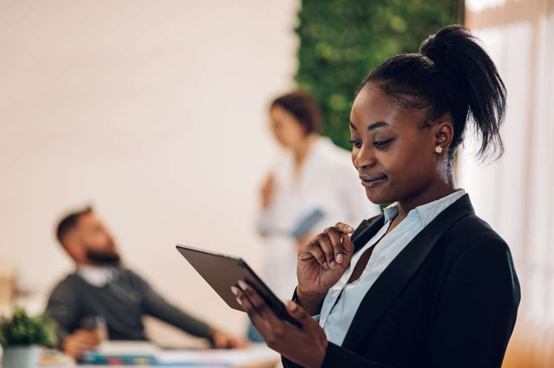 A Black professional woman smiling as she reads on her tablet in a bright office