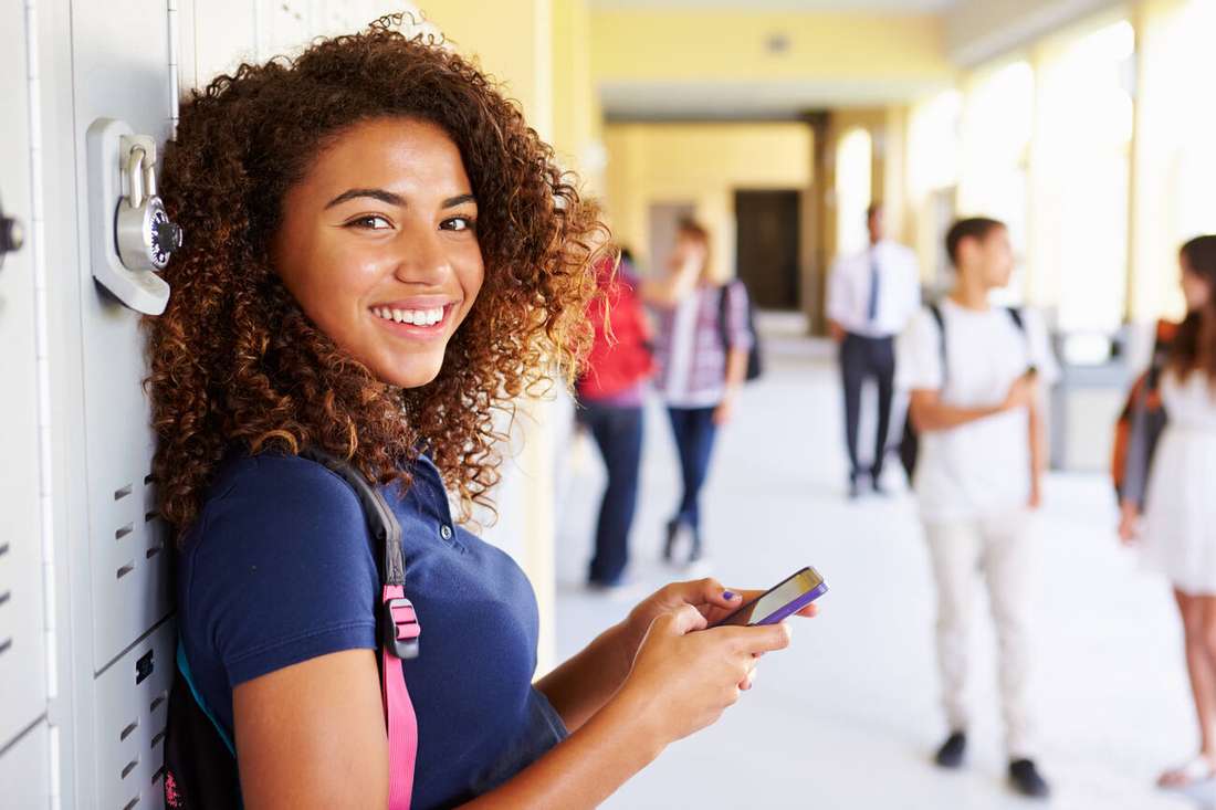 A smiling young woman leaning against a locker, holding her phone