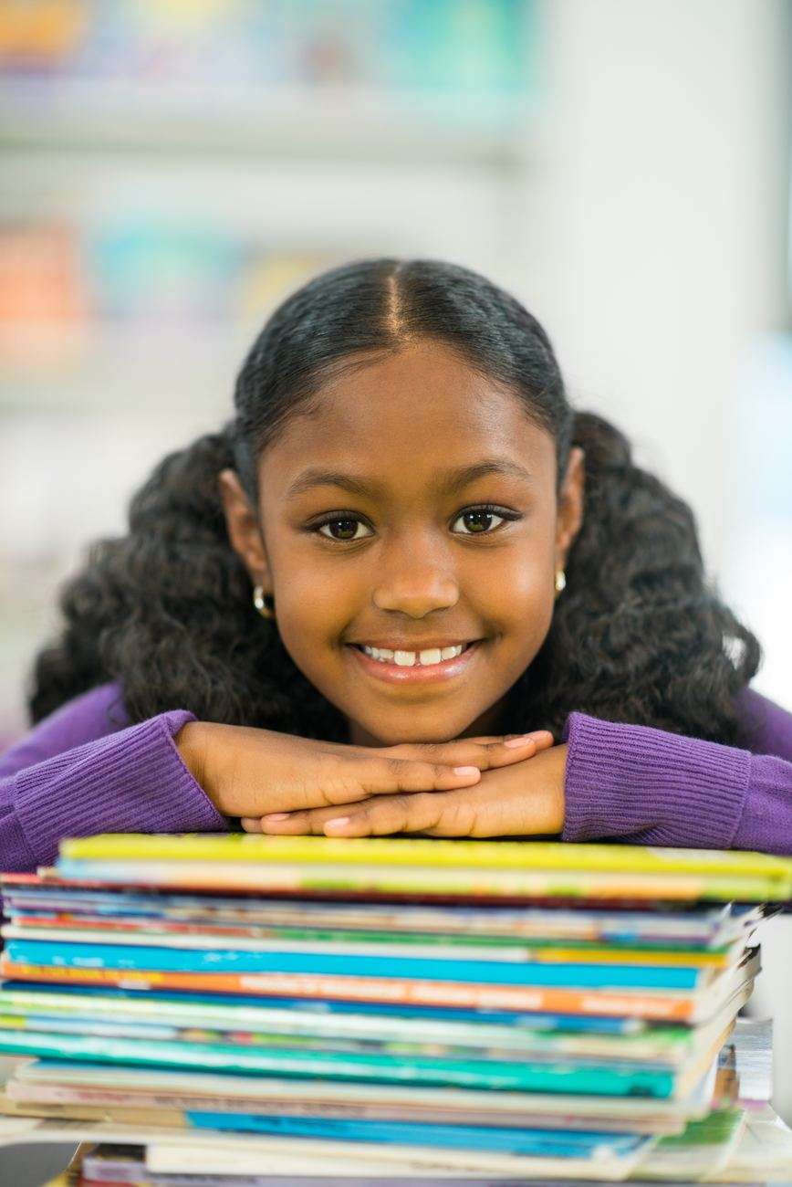 A smiling Black girl resting her chin on her hands on top of a tall stack of books