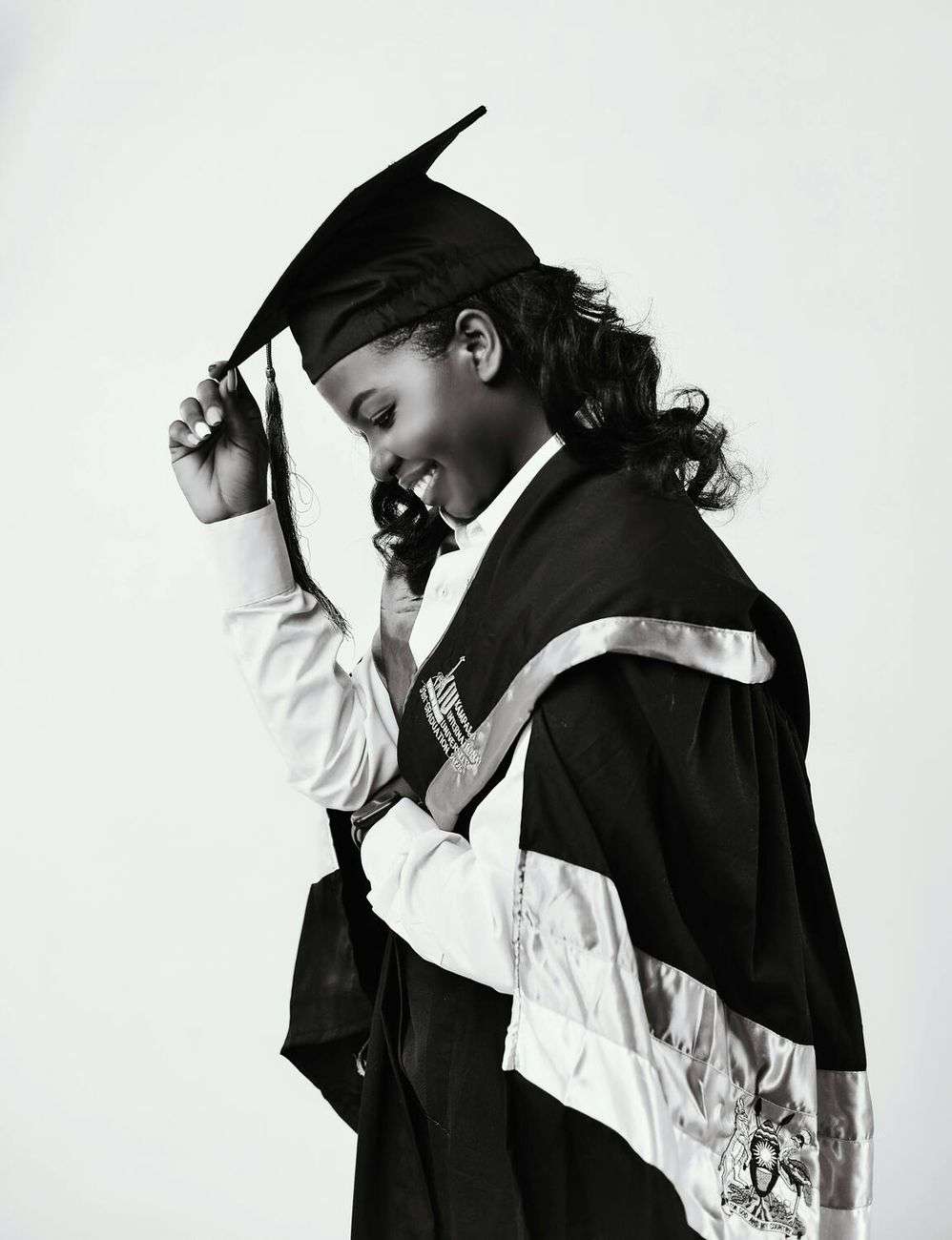 A smiling young Black woman in a graduation cap and gown, adjusting her tassel, in black and white