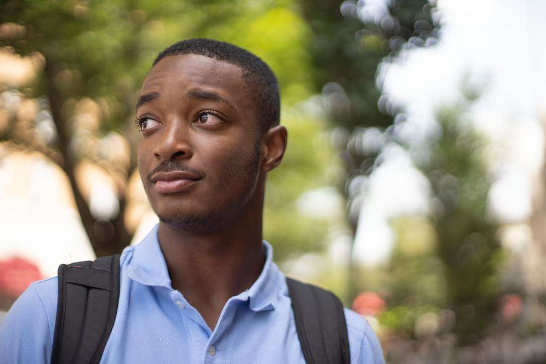 A young Black man with a backpack looking ahead thoughtfully, trees behind him