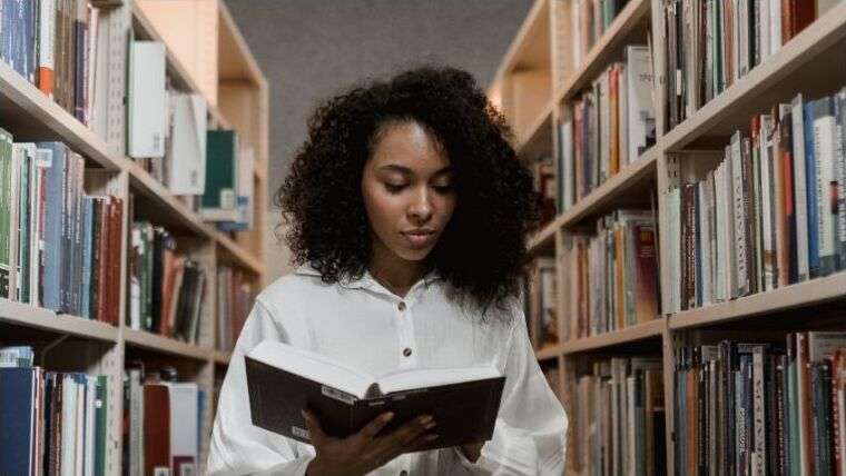 A young Black woman reading a book between library shelves