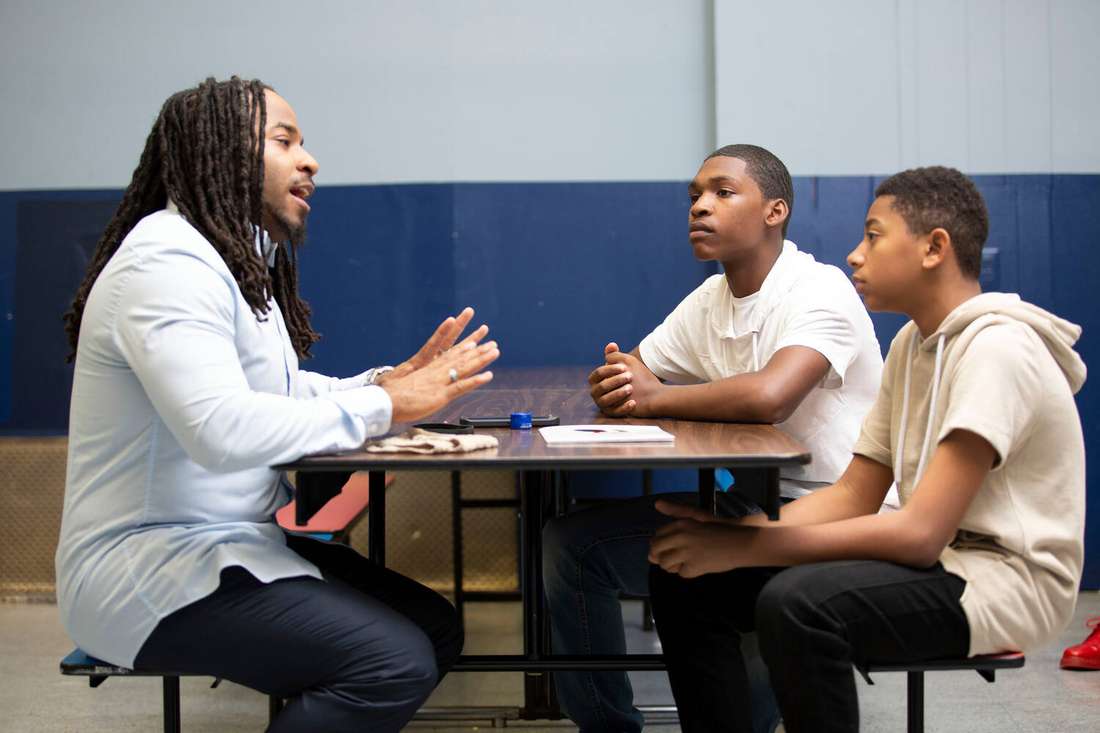 A Black male mentor talking with two teenage boys at a table