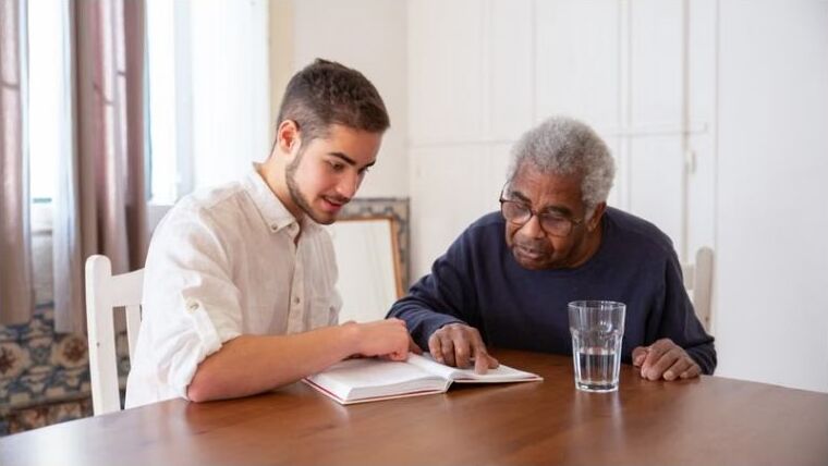 An elder and a young man reading a book together at a table