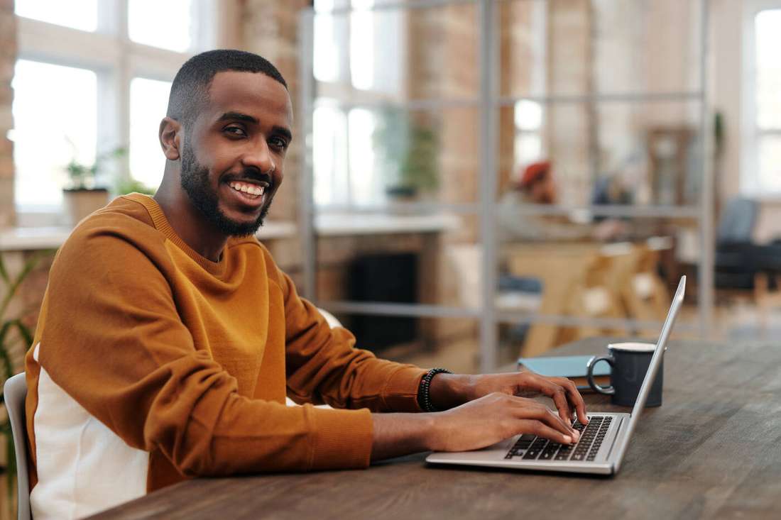A smiling man in an orange sweater working on his laptop